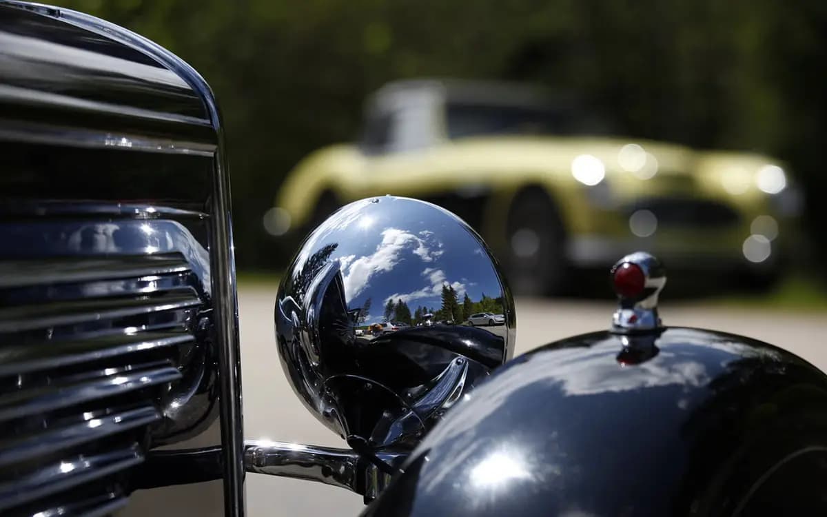 Close up of a classic car's glossy black fender and chrome headlight reflecting blue sky and trees